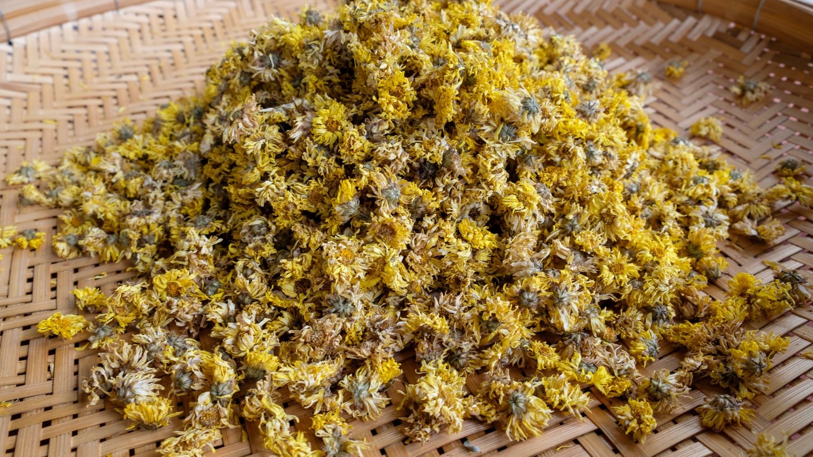 A pile of dried, golden-yellow blooms sits on a woven mat, with each flower head crinkled and curled.