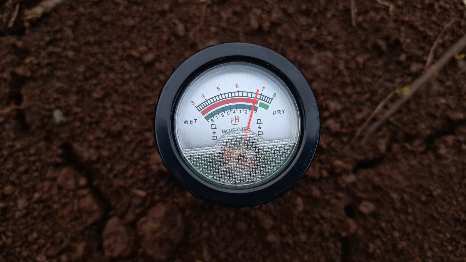 Close-up of a device inserted into soil to measure pH levels, featuring a digital display that shows moisture and pH readings, encased in a durable plastic body.
