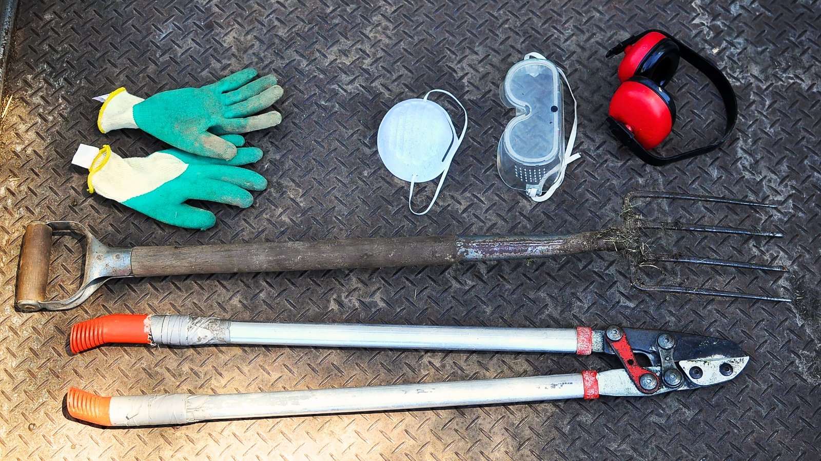 Garden tools including glove, mask, eyeglasses protector, earmuff, farm trident, compost fork, power pivot cut, and lopper are placed on a metal surface.